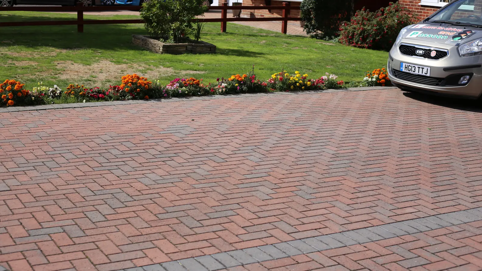 A block paved driveway running past a flower bed, with houses behind